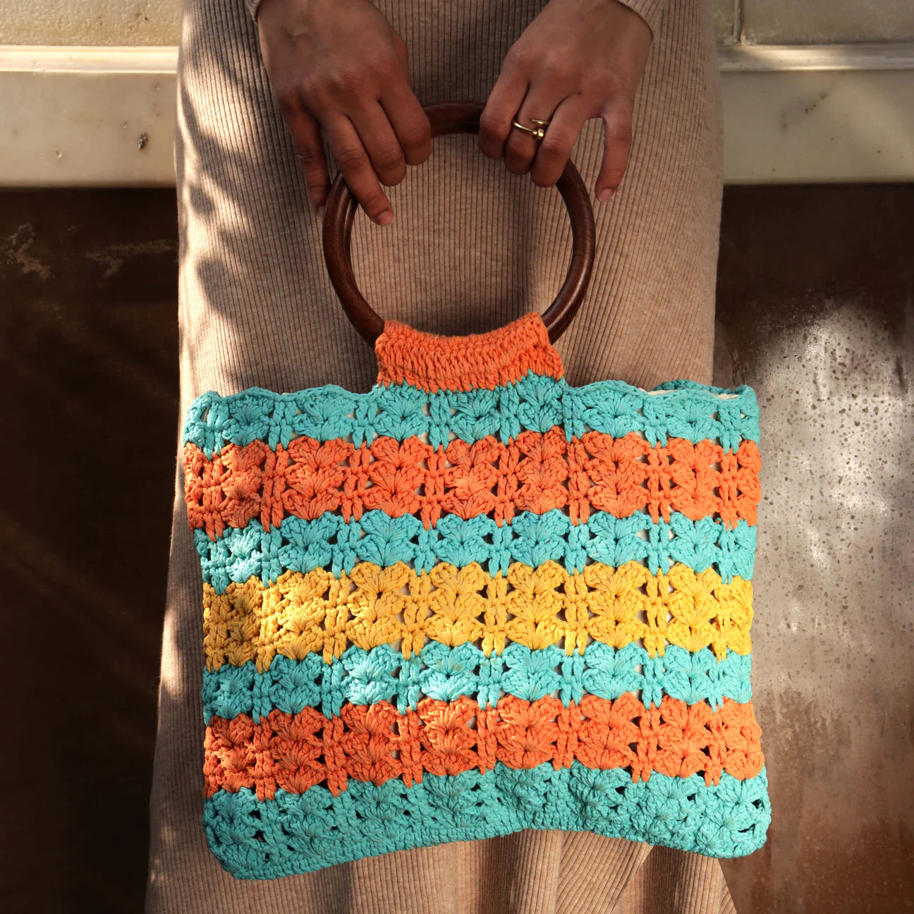 Upgrade your accessory game with our hand-crocheted rectangular handbag, featuring circular wooden handles, in eye-catching hues of orange, blue, and yellow for a stylish and vibrant appeal.